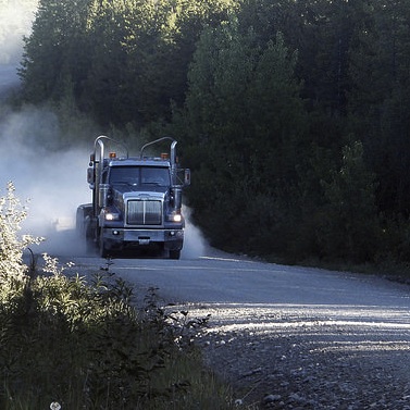 A logging truck coming down a logging road.  There are trees on both sides of the road and dust billowing behind the truck. 