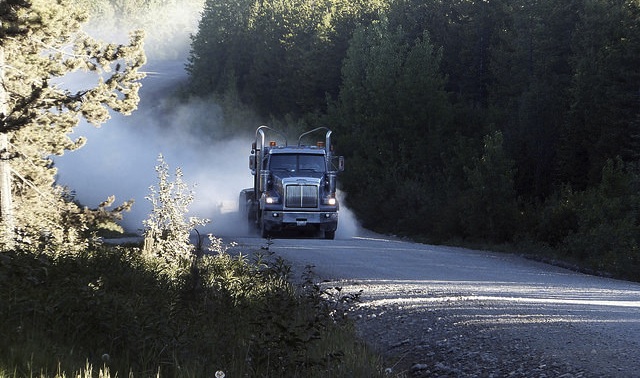 A logging truck coming down a logging road.  There are trees on both sides of the road and dust billowing behind the truck. 