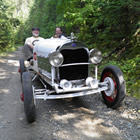 Photo of two older gentlemen sitting behind the wheel of a classic speedster car on a trail in the bush. 