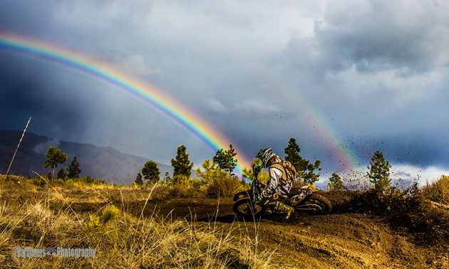 A dirt bike rider hits a berm right with a rainbow in the background. 