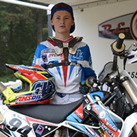 A young boy dressed in full moto gear standing by a race dirt bike. 