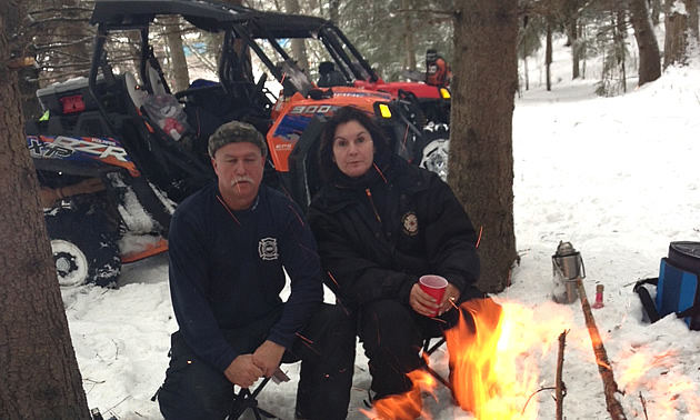 A man and woman kneeling down by a campfire in the wintertime. 