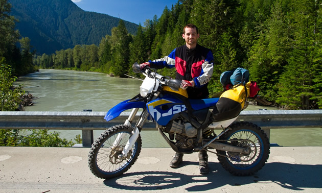 A dark haired man standing on a bridge with a dual sport bike.