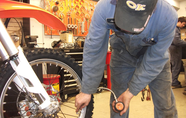 A man in a shop coat checking tire pressure on an orange dirt bike. 