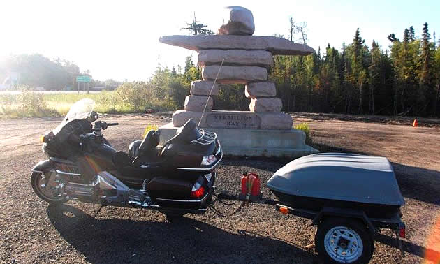 A statue of rocks stacked on each other is perched behind a motorcycle with a trailer. 