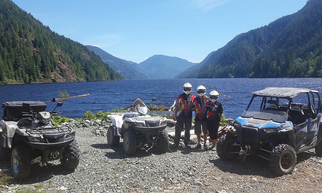 three atv riders standing beside a lake with mountains in the background