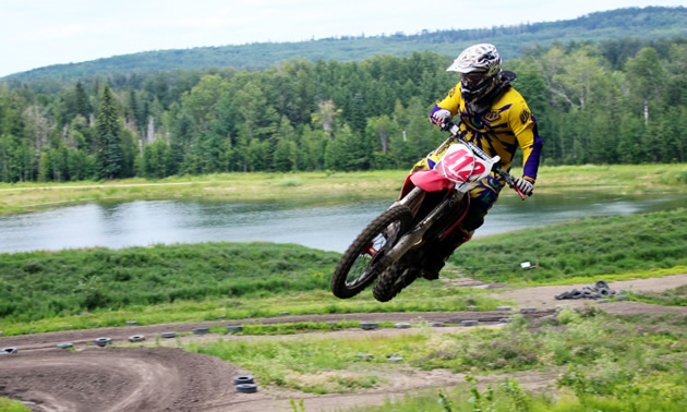 A motorcycle gets massive air in the foreground while a track and swampland is in the background.