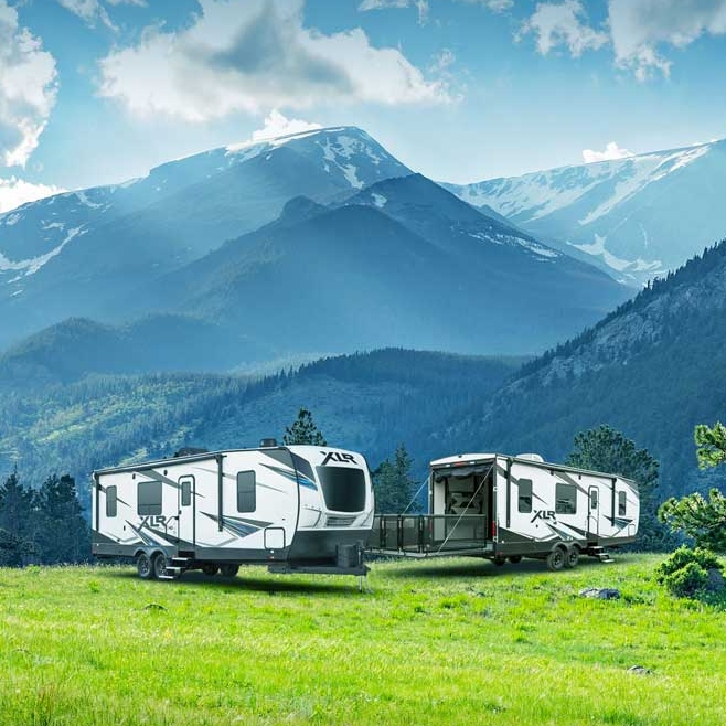 Two massive white RVs are parked with sun shining on the mountains in the distance