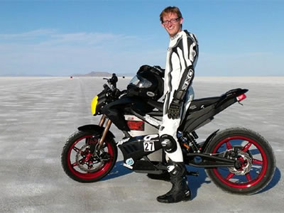 Photo of a young man dressed in white and black riding gear standing next to a black, red and white street bike. 